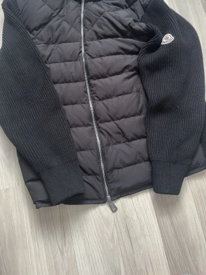 Svart cardiganjacka från Moncler - Snygg svart cardiganjacka från Moncler med quiltad front och stickade ärmar. Jackan har dragkedja framtill och en Moncler-logga på ena ärmen. Perfekt för dig som vill ha en stilren och exklusiv look.