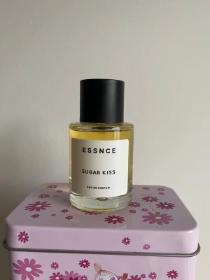 ESSNCE Sugar Kiss Eau de Parfum - ESSNCE Sugar Kiss Eau de Parfum i en rund glasflaska med svart lock och enkel vit etikett. Doften har en ljusgul vätska och flaskan har en stilren, modern design som känns både trendig och minimalistisk.