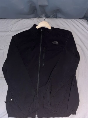 Svart vindjacka från The North Face - Snygg svart vindjacka från The North Face med dragkedja framtill och diskret logga på bröstet och ryggen. Jackan har hög krage, två sidofickor med dragkedja och är tillverkad i ett lätt, vindtätt material. Perfekt för blåsiga dagar.