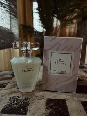 Avon Rare Pearls edp, 50ml - Rare Pearls Eau de Parfum från Avon i en elegant, mjölkvit glasflaska med facetterad design och genomskinlig dekorativ kork. Doften kommer i originalkartong med stilren, ljuslila färg och subtilt mönster. Perfekt för dig som gillar exklusiva och feminina parfymer.