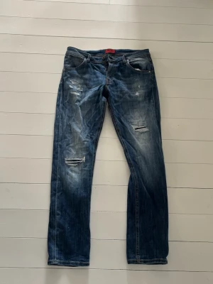 Jack and Jones jeans  - Säljer ett par blå jeans med raka ben och flera slitningar och hål på framsidan. Jeansen har klassisk femficksdesign och knäppning fram. Snyggt tvättade detaljer och en avslappnad look som passar till det mesta. Hör av dig vid frågor mvh Malte 