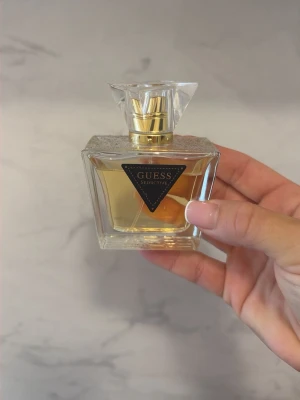 Guess Seductive parfym - Guess Seductive parfym i en elegant, fyrkantig glasflaska med gulddetaljer och genomskinlig, kristalliknande kork. Doften har en lyxig känsla och flaskan har en snygg svart etikett med guldtext framtill.