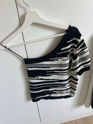 Randig offshoulder stickad topp - Snygg svart och vit stickad topp med offshoulder-design. Toppen har ett unikt randigt mönster och är croppad i modellen. Toppen är från Lindex och är i storlek XS
