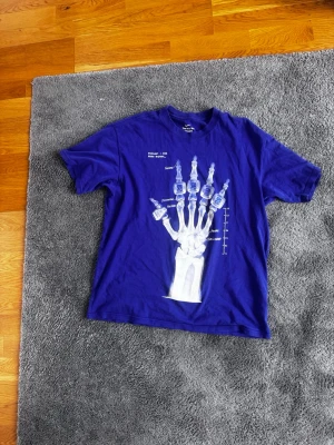 Blå kobe, Nike t-shirt - Cool blå t-shirt från Nike med ett stort vitt röntgenmotiv av en hand på framsidan och Nike-logga på baksidan. T-shirten har korta ärmar med baggy/ oversized fit. Perfekt för dig som gillar unika tryck och sportig stil.