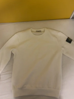 Vit sweatshirt från Stone Island - Vit sweatshirt från Stone Island med klassisk rund halsringning och ribbade muddar. Tröjan har den ikoniska Stone Island-loggan på vänster ärm. Perfekt för en clean och stilren look.