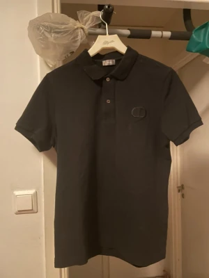 Svart pikétröja från Dior - Säljer en svart pikétröja från Dior med klassisk krage och knappar framtill. Tröjan har korta ärmar och en diskret Dior-logga broderad på bröstet. Perfekt för en clean och stilren look.