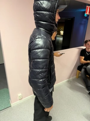 Svart dunjacka med huva - Snygg svart dunjacka med glansig finish och quiltad design. Jackan har dragkedja framtill, två sidofickor och en justerbar huva. Perfekt för kalla vinterdagar och ger en modern look.