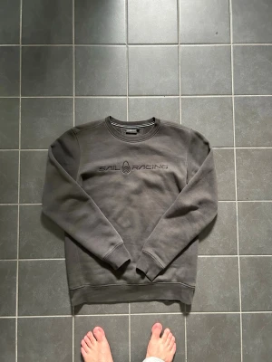 Mörkgrå Sail Racing sweatshirt - Mörkgrå sweatshirt från Sail Racing med rund halsringning och diskret logga broderad på bröstet. Tröjan har långa ärmar, ribbade muddar vid ärmslut och nederkant samt en klassisk passform. Perfekt för en avslappnad och stilren look.
