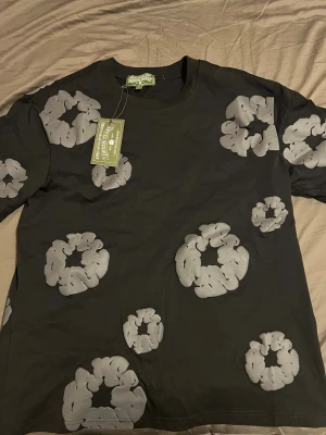 Denim tears t-shirt - Säljer en svart t-shirt från Denim Tears med stora gråa blommönster över hela tröjan. Klassisk rund hals och korta ärmar. Perfekt för dig som vill sticka ut med en unik och trendig look.