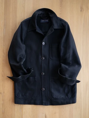 A Day’s March Chore Jacket, size M - Stilren svart overshirt i kraftigt bomullstyg med tre stora utanpåliggande fickor och knäppning framtill. Klassisk krage och raka ärmar som kan vikas upp för en avslappnad look. Perfekt lager på lager-plagg för en trendig streetwear-stil.