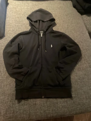 Svart hoodie från Polo Ralph Lauren - Snygg svart hoodie från Polo Ralph Lauren med dragkedja framtill och klassisk logga broderad på bröstet. Hoodien har huva med dragsko och två fickor framtill. Perfekt för en chill och stilren look.