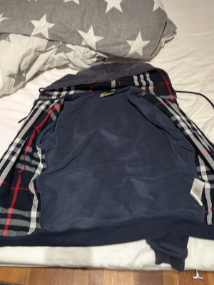 Mörkblå hoodie med dragkedja - Snygg mörkblå hoodie med dragkedja och huva. Insidan har ett klassiskt rutigt mönster i rött, vitt och svart. På bröstet finns en diskret broderad logga. Hoodien har ribbade muddar och känns mjuk och bekväm, perfekt för en avslappnad stil.