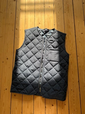 Svart quiltad dunväst med dragkedja - Stilren svart dunväst med quiltat mönster och dragkedja framtill. Västen har en bröstficka och är ärmlös med rundad hals. Perfekt för lager-på-lager och ger en clean look till din outfit.