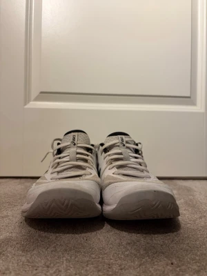 Vita Asics Gel-Tactic 12 innebandyskor - Säljer ett par vita Asics Gel-Tactic 12 innebandyskor med svarta detaljer och snörning. Skorna har en slitstark sula och ventilerande meshmaterial på ovandelen. Perfekta för sporter som innebandy.