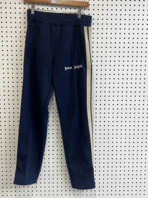 Blåa Palm Angels mjukisbyxor S - Säljer ett par mörkblå mjukisbyxor från Palm Angels med vita sidoränder och logga på vänster ben. Byxorna har resår i midjan och raka ben. Tillverkade i polyester för en skön och sportig känsla.