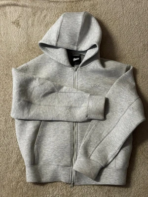 Grå zip hoodie från Stussy - Snygg grå zip hoodie från Stussy med broderad logga på bröstet. Tillverkad i mjukt material med borstad insida och två fickor framtill. Klassisk huva och ribbade muddar vid ärmar och nederkant. Perfekt för en chill och stilren look.