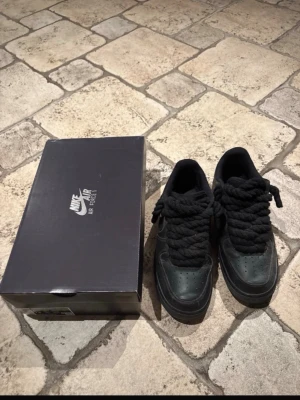 Nike Air Force 1 med tjocka skosnören - Säljer ett par svarta Nike Air Force 1 sneakers med unika, extra tjocka svarta skosnören. Skorna är låga och har klassisk perforerad tåbox och Nike-logga på sidan. Tillverkade i läder med textildetaljer. Boxen medföljer. Pris kan förhandlas.