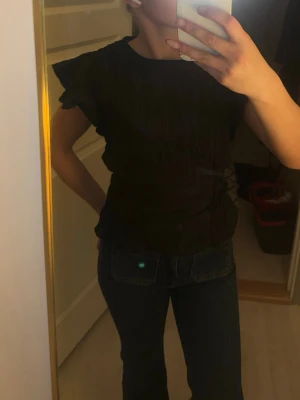 Svart topp med volangärmar - Säljer en svart topp med korta volangärmar och rund halsringning. Toppen har en smockad struktur som ger en snygg passform och lite extra detaljer. Perfekt att styla med jeans eller kjol för en trendig look.