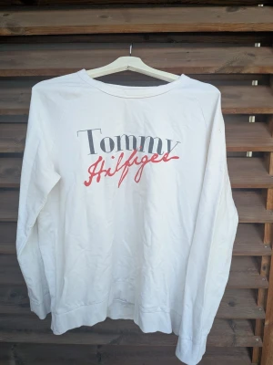 Vit sweatshirt Tommy Hilfiger - Vit sweatshirt från Tommy Hilfiger med tryckt logga i svart och rött framtill. Tröjan har rund halsringning och långa ärmar. Den har en liten fläck på ena axeln, går säkert att bleka bort men har inget fläckborttagningsmedel hemma så har ej försökt.