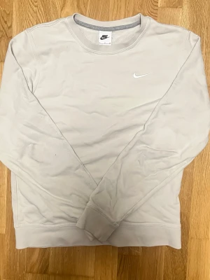 Långärmad tröja från Nike - En stilren beige långärmad tröja från Nike med klassisk rund halsringning och diskret broderad vit Nike-logga på bröstet. Tröjan har långa ärmar och ribbade muddar vid ärmslut och nederkant. Perfekt för en avslappnad och sportig look.