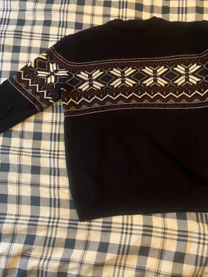 Svart stickad tröja med snöflingemönster - Mysig svart stickad tröja med klassiskt snöflingemönster i vitt och vinrött över bröstet och ärmarna. Tröjan har rund halsringning och ribbade muddar vid ärmslut och nederkant. Perfekt för kalla dagar och vintersäsongen.