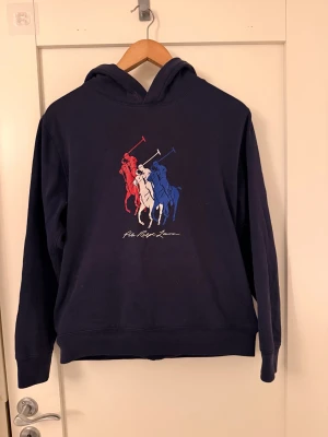 Mörkblå hoodie med polo-tryck - Snygg mörkblå hoodie från Polo Ralph Lauren med stort tryck av tre polospelare i rött, vitt och blått på bröstet. Klassisk huva och ribbade muddar. Perfekt för dig som gillar streetwear och vill ha en stilren men ändå sportig look. Den är barn xl men skulle säga herr xs 