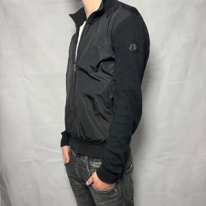 Moncler Cardigan  - Snygg svart Moncler Cardigan med nylon front och stickade ärmar. Koftan har ribbade muddar, hög krage och två dragkedjeförsedda fickor framtill. Perfekt för dig som gillar en sportig men ändå stilren look.