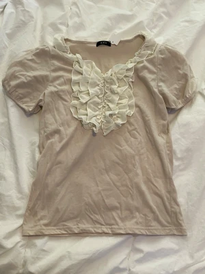 Beige topp med volangdetaljer - Söt beige topp med korta puffärmar och volangdetaljer framtill samt runt halsringningen. Köpt på secondhand i Japan