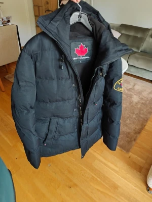 Svart dunjacka från Zavetti Canada mycket bra skick storlek L  - Säljer en svart dunjacka från Zavetti Canada med huva och dragkedja. Jackan har två stora fickor framtill och en patch med logga på ena ärmen. Perfekt för kalla vinterdagar och riktigt skön att bära.