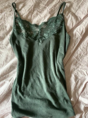 Grön ribbad linne med spets - Snygg grön linne med tunna axelband och ribbad struktur. Överdelen har vacker spetsdetalj i samma färg som ger en feminin touch. Perfekt att styla under en skjorta eller kavaj. Materialet känns mjukt och följsamt mot huden.