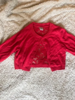 Röd spetskofta från Lindex - Snygg röd kofta från Lindex med spetsdetaljer på ryggen och knappar framtill. Koftan har rund halsringning, ribbade muddar och är tillverkad i ett mjukt material. Perfekt för att lyfta en outfit med lite extra färg och stil.