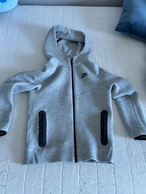 Grå Nike hoodie med dragkedja - Snygg grå hoodie från Nike med hel dragkedja framtill och två svarta dragkedjeförsedda fickor. Hoodien har en klassisk huva och diskret Nike-logga på bröstet. Tillverkad i mjukt och bekvämt material, perfekt för en avslappnad stil.