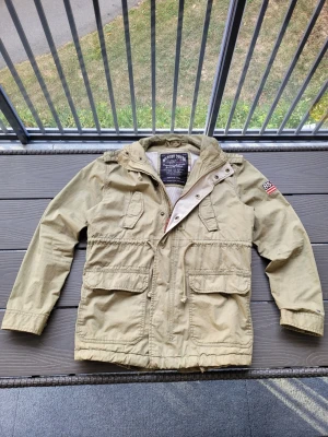 Grön tommy hilfiger jacka - Stilren grön jacka med huva och flera praktiska fickor framtill. Jackan har dragsko i midjan och en patch med siffran 85 på ena ärmen. Tillverkad i slitstarkt bomullsmaterial och har knapp- och dragkedjestängning. Obs! Jackan har mindre tecken på användning och även mindre fläckar längst ned på ärmarna(väldigt små). Dessa syns dock ej vid användning och går lätt att ta bort.    Den ser även något mörkare ut i verkligheten(lite mer oliv färgad).