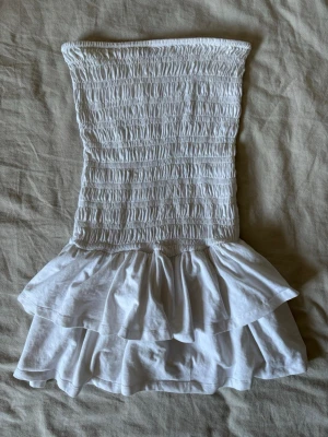 Vit volangklänning från Gina Tricot - Säljer en söt vit kort klänning från Gina Tricot med smockad överdel och dubbla volanger nedtill. Klänningen är axelbandslös och har en figurnära passform upptill. Perfekt för varma dagar eller sommarens alla tillfällen!
