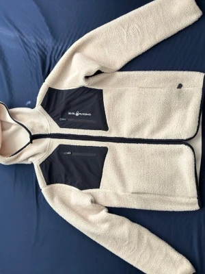 Beige fleecejacka från SailRacing - Säljer en beige fleecejacka från Sail Racing med svarta bröstfickor och dragkedja. Jackan har hög krage, hel dragkedja framtill och två bröstfickor med dragkedja. Perfekt för lager-på-lager och outdoor vibes.