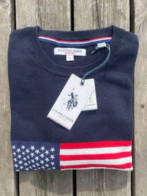 Marinblå stickad tröja U.S. Polo Assn. - Snygg marinblå stickad tröja från U.S. Polo Assn. med amerikanska flaggan broderad på bröstet och logotyp under. Rund halsringning och ribbade muddar. Perfekt för dig som gillar klassisk och sportig stil.