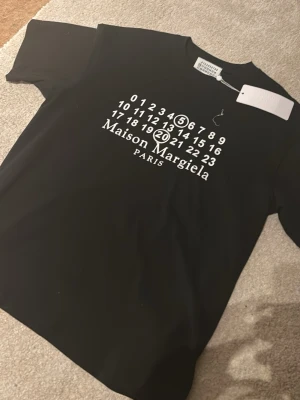 Maison Margiela svart t-shirt - Svart t-shirt från Maison Margiela med ikoniskt vitt tryck framtill som visar märkets logotyp och siffror. Klassisk rak passform och rund halsringning. Perfekt för dig som gillar stilrena och trendiga plagg.