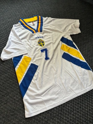 Svensk landslagströja Adidas nr 7 Larsson - Vit landslagströja från Adidas med blå och gula detaljer, nummer 7 på framsidan och svensk landslagssköld. V-ringad krage med blå, gul och röd rand. Perfekt för dig som vill stötta Sverige på match eller träning.