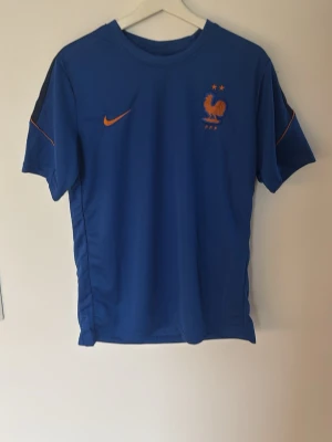  Nike Frankrike fotbollsett - Säljer en blå fotbollströja från Nike med Frankrikes landslagsemblem och två stjärnor på bröstet. Orange Nike-logga och FRANCE tryckt i orange på ryggen. Tröjan har korta ärmar och svarta detaljer på axlarna. Perfekt för träning eller match.