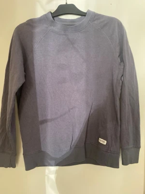 Lila sweatshirt med rund hals - En enkel och stilren gråblå sweatshirt med rund hals och långa ärmar. Tröjan har ribbade muddar vid ärmslut och nederkant samt en liten etikett nertill. Perfekt för en avslappnad look.