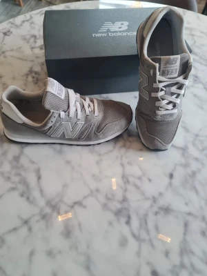 Grå New Balance 373 sneakers - Snygga grå New Balance 373 sneakers med klassisk design och vita detaljer. Skorna har mesh och mocka på ovansidan, vit sula och vita skosnören. Perfekta för dig som gillar stilrena och sportiga sneakers.
