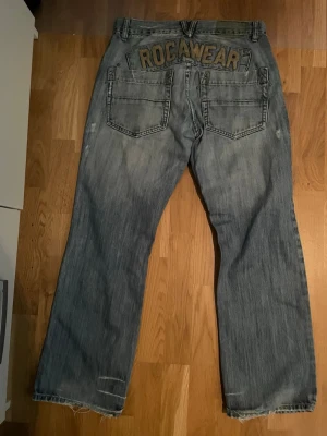 Bootcut jeans från Rocawear - Säljer ett par ljusblå bootcut jeans från Rocawear med slitna detaljer och bred passform. Jeansen har stora bakfickor med broderad logga och klassisk femficksdesign. 
