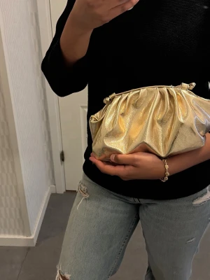 Guldig kuvertväska med veckad design - Säljer en snygg kuvertväska i metallic guld med veckad och mjuk form. Väskan har en modern look och är tillverkad i glansigt material som verkligen sticker ut. Perfekt accessoar för att lyfta din outfit. Kommer med band❤️