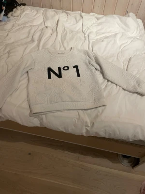Vit sweatshirt med text N°1 - Kan förhandla i snabb afär. Kommer från ett djurfritt hem.Säljer en vit sweatshirt med upphöjt mönster och svart text 'N°1' på bröstet. Tröjan har rund halsringning och långa ärmar.