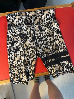 Leopardmönstrade cykelshorts Stronger - Snygga cykelshorts från Stronger med leopardmönster i beige, svart och brunt. Hög midja och två svarta ränder på ena benet. Tillverkade i 85% återvunnen polyester och 15% elastan för stretch och komfort. Använda fåtal gånger, säljer flera stonger byxor på min sida så in o kika!