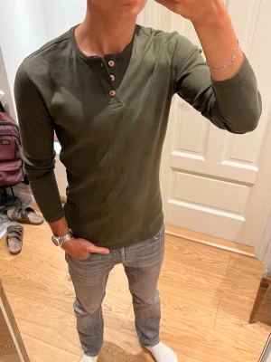Olivgrön henleytröja - Snygg olivgrön långärmad henleytröja med ribbad struktur och tre träknappar framtill. Tröjan har en skön slim passform. Använd 1-2 gånger, alltså inga defekter. Skriv om du undrar något!