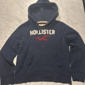 Hollister hoodie - Mörkblå hoodie från Hollister med broderad vit logotyp och röd fågel på bröstet. Klassisk känguruficka framtill och huva med mjukt foder. Perfekt för dig som gillar en avslappnad och sportig stil.