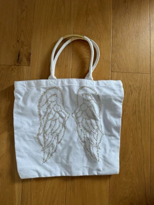 Vit väska med änglavingar - Snygg vit väska från Victoria's Secret med glittrande änglavingar i guld på framsidan. Väskan har dubbla handtag med gulddetalj och är gjord i ett mjukt tygmaterial. Perfekt för att bära allt du behöver till skolan eller stan.