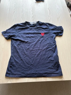 Comme des garcons T-shirt  - Snygg svart t-shirt från comme de garcons med ett ikoniskt rött hjärta med ögon broderat på bröstet. Klassisk rund hals och korta ärmar. Perfekt för dig som gillar stilrena plagg med en twist.