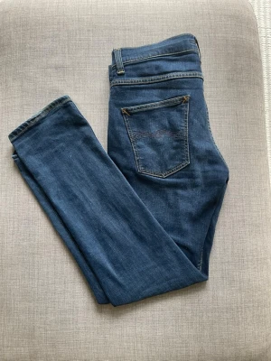 Blå jeans från Nudie Jeans 32/32 - Säljer ett par klassiska blå jeans från Nudie Jeans i storlek 32/32. Jeansen har en smal passform, fem fickor och snygga kontrastsömmar. Tillverkade i mjukt denimtyg som sitter skönt hela dagen. Perfekta till din vardagsstil!